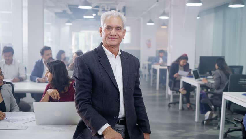 Movement of a Smiling happy Indian Asian senior male corporate businessman with arms crossed and looking at the camera confidently while busy office employees working in the back in a modern office | Shutterstock HD Video #1107004477