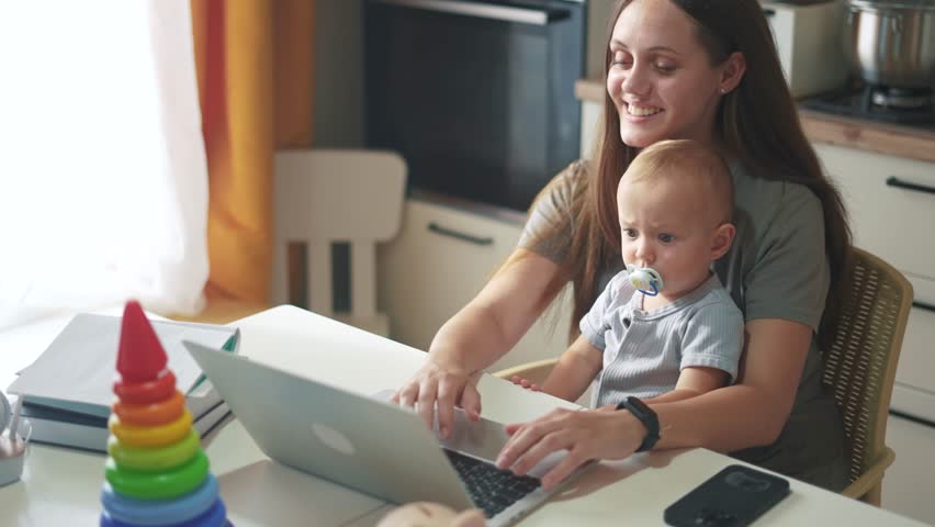 mother with a child sits at a table and works on a laptop. remote work mom baby concept. a woman works in a family setting on the table are her lifestyle child's toys. computer work Royalty-Free Stock Footage #1107007901