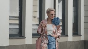 Fashionable mature blonde woman with short hair in trendy clothes pink floral cardigan, cargo jeans, sunglasses walking outside on street. Fashion street style