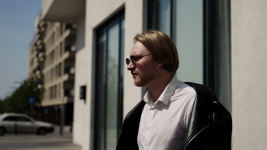 A brutal man walks along the building and puts on a black jacket.