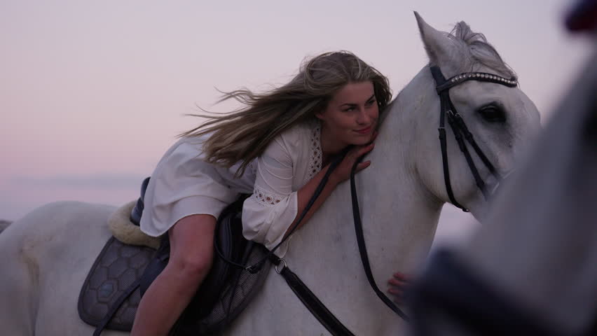 A medium shot of a long-haired girl hugging and caressing her white horse during sunset
