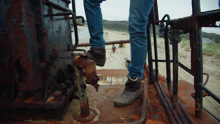 A young man with blue jeans and brown shoes climbs up the stairs of the old rusty ship