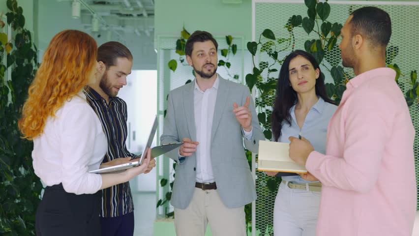 Handsome Guy Team Leader Boss Speaking about Project with his Colleagues in the Standup Meeting Standing in Office. Positive Multiracial Team of Managers Working Together in Office