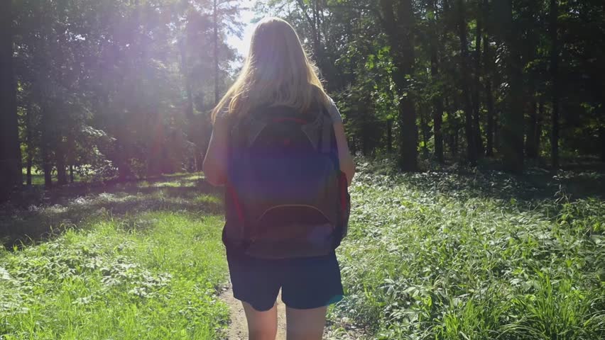 Girl with backpack on her back walks along path in forest. Short shorts, white T-shirt and large backpack. Rays of sun into lens, grass, trees and atmospheric color correction create pleasant feeling
