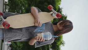 Girl with a longboard in her hands standing against the background of trees and drinking coffee. Vertical video