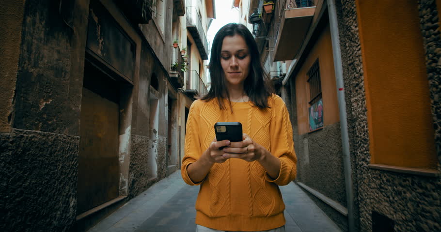 Attractive cheerful woman walk on old city street with mobile phone chatting with friends or using it for navigation 