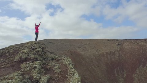 AERIAL: Flying around cheerful young woman standing on the edge of big volcano crater with hands raised
