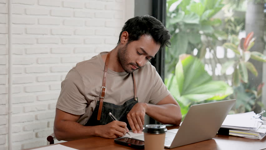 Handsome Indian man is a barista and owner of small business cafe. sitting at the table take your mind seriously to prepare to order raw materials of the coffee shop via online with notebook computer Royalty-Free Stock Footage #1107063067