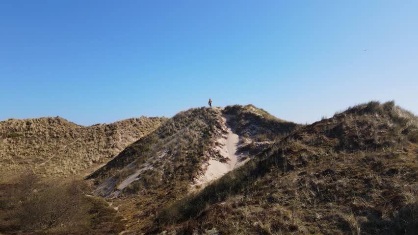 A drone flies towards a lone man perched on top of a dune, his bright yellow jacket standing out against the sky’s deep blue hue, as a cozy village near the sea is revealed.