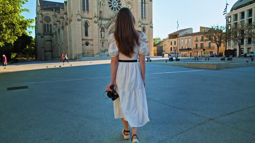 A girl tourist visits catholic church in the neo-Gothic style in Nimes, France. Young woman with a camera explores a historical building with two bell towers on the facade surmounted by high Spiers.