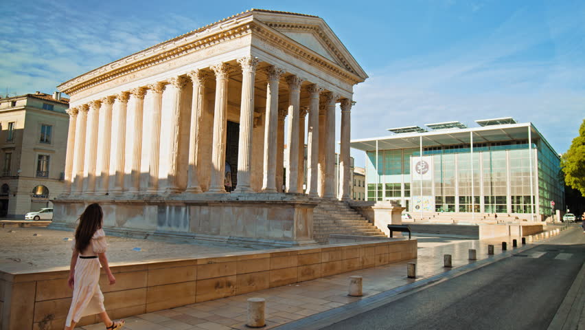 Young woman explores The Roman temple in Nimes, Occitanie, France on a sunny summer day. Beautiful girl in a dress looks at the Antique temple decorated with columns and friezes in France.
