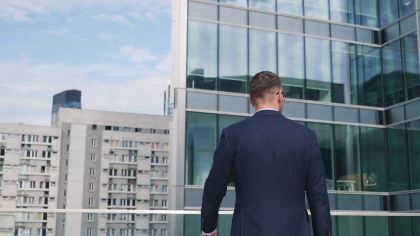 Relaxed male CEO businessman in suit standing on rooftop glass terrace in office building overlooking modern city downtown with high-rise skyscrapers on sunny day. Business people, gimbal slow motion Royalty-Free Stock Footage #1107072573