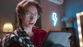 Asian guy in glasses with tablet sits on sofa, online communication. Vivid expressive emotions. Cinematic advertising. Expresses joy, he got good news. Concept of modern virtual lifestyle