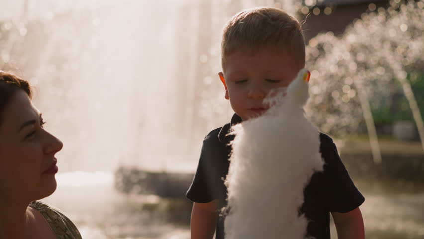 Happy mother looks at little son biting sugar cotton wool near sister against fountain spraying jets closeup slow motion. Woman with children in park Royalty-Free Stock Footage #1107082197