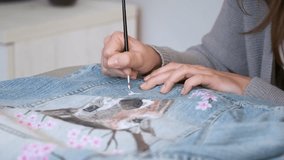 Unrecognizable woman painting a design of a deer and flowers in a denim jacket. Woman's creative expression through painted clothing designs.