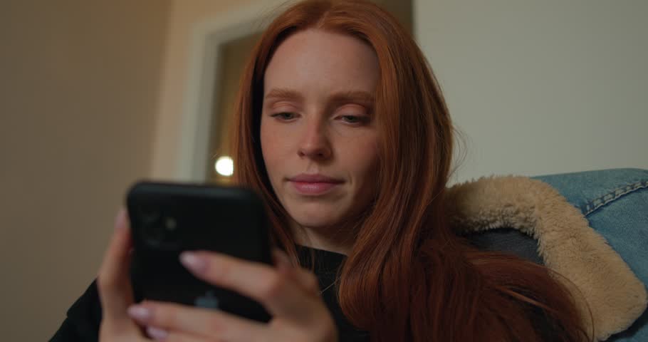 young red-haired woman in casual clothes watching online vebinar, studing online slow motion, girl chat with friends, conversation, , read online content, relax at home, rest in chair, use online app