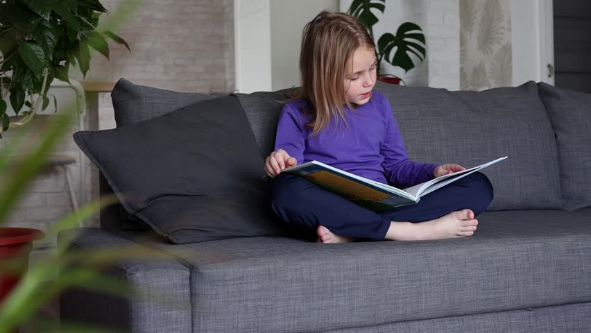 a little blonde girl is reading a big book sitting on the sofa in the living room.Smile and interest in reading.Self-education of children, reading literature at home, interesting leisure. Royalty-Free Stock Footage #1107091827