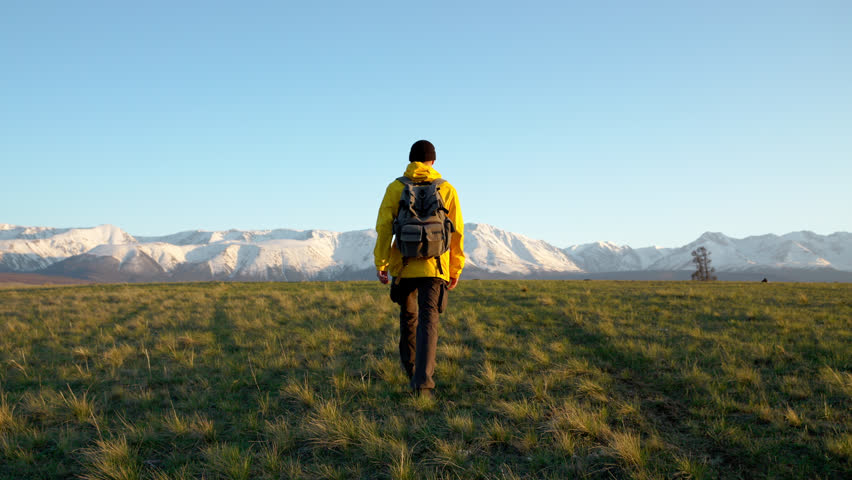 Young hiker man trekking along wild cliff, going way forward to mountains at sunset. Extreme travel destinations, walk path on high area alone