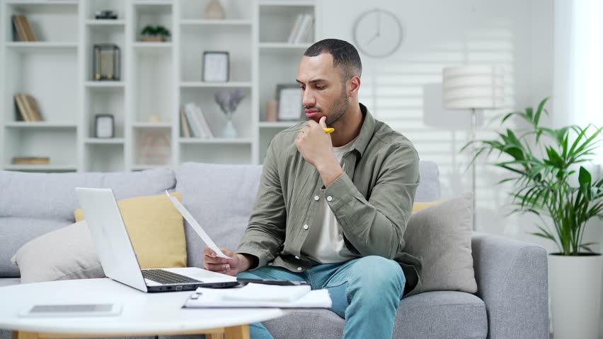 Thoughtful man calculates household finances and accounts sitting at home in the living room using a laptop computer and calculator. Male businessman doing a paperwork job bils receipts finances Royalty-Free Stock Footage #1107096331