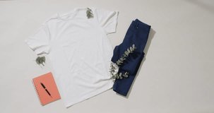 Video of flat lay of white t shirt, denim trousers, notebook and copy space on white background. Clothing, fabric, texture and materials concept.