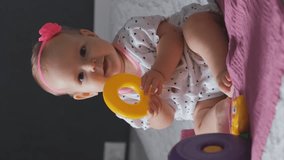 Vertical video. Cute baby girl playing toys while sitting on bed at home, early development for infant