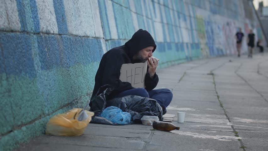  homeless poor young man sitting on the ground in the city with a Help table while asking for money to survive. Eating old unhealthy food