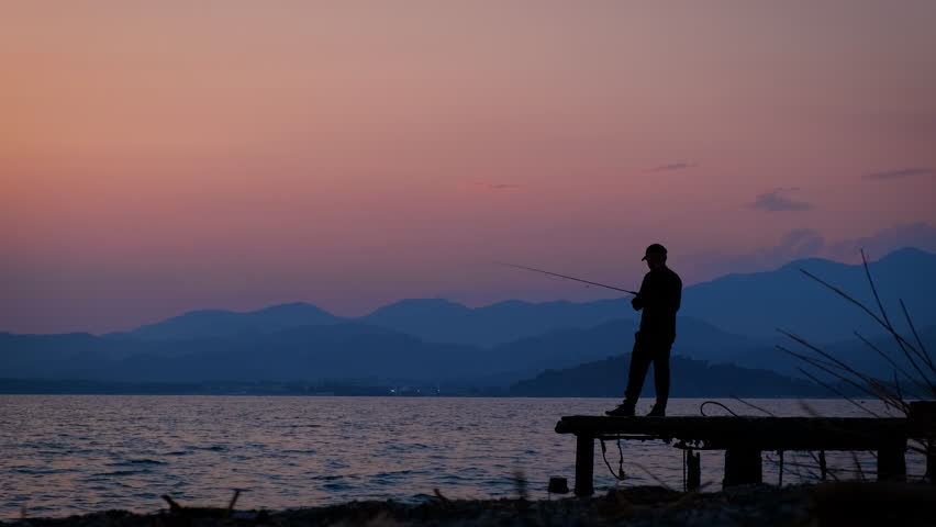Male silhouette hold fishing rod in evening. A view of man silhouette stay on the small wooden pier and hold a fishing rob in her hands against pink and blue sky. Royalty-Free Stock Footage #1107103393