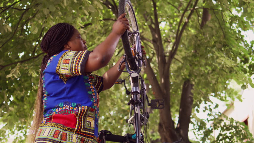 Energetic black woman grasping and removing broken bicycle wheel for further maintenance outside. Athletic african american lady inspecting bike components for damages to repair with work tool. | Shutterstock HD Video #1107103843