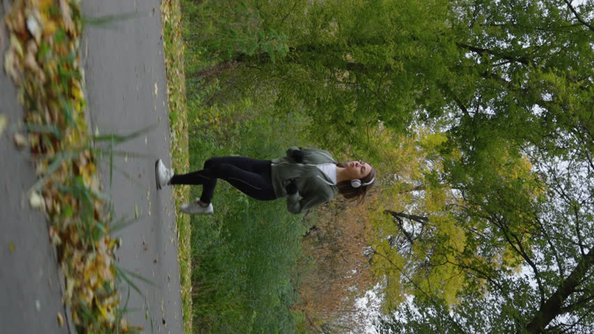 Vertical Screen: Slow motion woman wearing sportswear, gloves and headphones jogging on road against forest, fallen leaves lying on ground. Active female exercising on chilly autumn morning