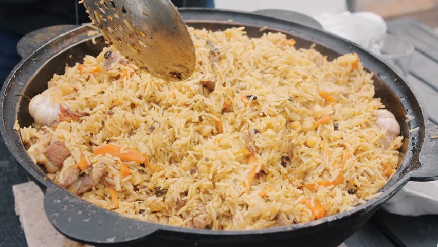 Cook puts prepared pilaf into guest bowl using slotted spoon. People ready to taste delicious dish with rice and vegetables at picnic closeup
