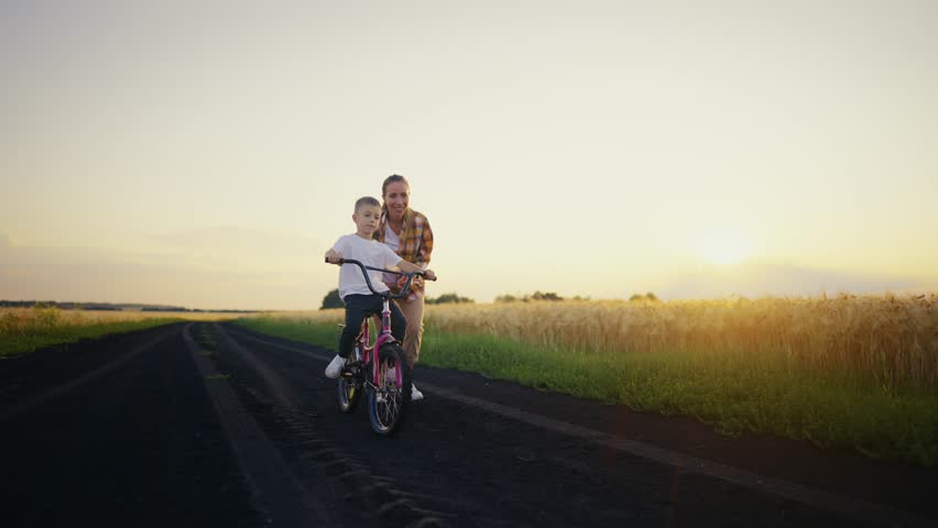 Mom holding bike with sitting on it son teaching him to ride in village on summer vacation at sunset. Mother running after bicycle. Happy family having outdoors activity on weekend, summer vacations. Royalty-Free Stock Footage #1107122979