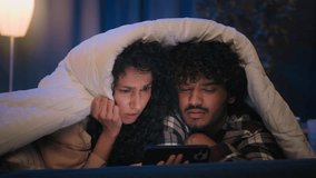 Gadget internet addicted multiracial couple happy family man woman boyfriend girlfriend at night at home under blanket watching disgust bad news video horror movie with mobile phone confused puzzled - Powered by Shutterstock - Get 15% off with code: PIKWIZARD15