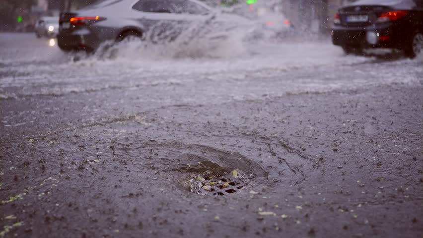Slow motion, Swift stream of rainwater flows into storm drain, cars are driving at city crossroads on background. Heavy rain leads to sewage overflow, flood in city