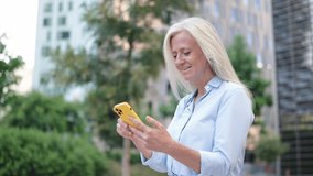 Grey-haired professional woman enjoys social clips among city greenery and skyscrapers, smiles and surprise faces
