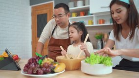 Happy Asian family with father, mother, and daughter enjoy learning how to make a salad video in the internet on tablet together. Asian family enjoy making a vegetable salad together.