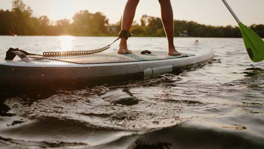 Close up of active young woman standing on sup board with safety rope tied on leg and paddling among calm lake. Summer activity on water of african american sports lady during beautiful sunset.