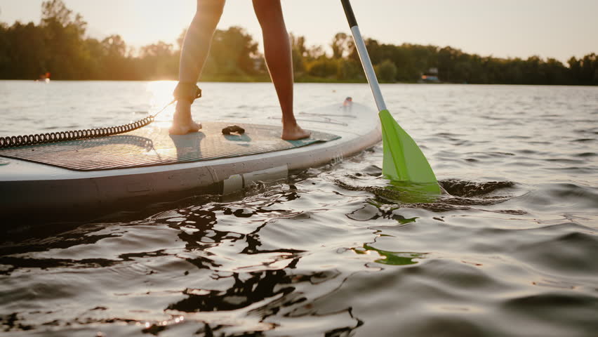 Close up of active young woman standing on sup board with safety rope tied on leg and paddling among calm lake. Summer activity on water of african american sports lady during beautiful sunset.