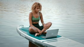 Beautiful multicultural woman with sporty body shape having video call on wireless laptop while balancing on sup board among city lake. Concept of people, technology and healthy lifestyles.