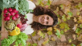 Happy mature woman holding crate full of fresh vegetables, looking at camera and smiling, posing outdoors by stone wall with yellow ivy leaves in autumn garden. Video portrait, vertical clip - Powered by Shutterstock - Get 15% off with code: PIKWIZARD15