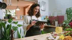 Mid-aged woman in apron standing by kitchen table with food ingredients, using digital tablet and searching for video recipes on the Internet before cooking dinner at home