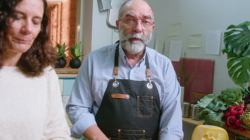 Mature woman standing next to husband cutting cheese, helping him to cook dish and commenting on camera during online culinary masterclass or food vlog