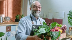 Senior chef with gray hair and beard holding a wooden crate full of fresh vegetables, smiling and posing for the camera in the kitchen. Medium shot, video portrait