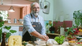 Senior male food blogger in apron cracking eggs in glass bowl, whisking and commenting on camera, filming video recipe or cooking class in the kitchen