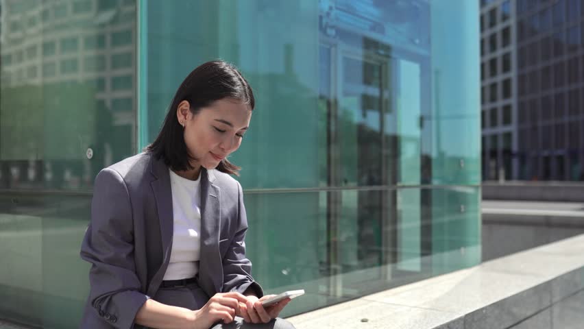 Smiling young Asian business woman holding smartphone using mobile apps tech, happy professional chinese businesswoman making online payment on cell phone reading news sitting on busy big city street. Royalty-Free Stock Footage #1107145423