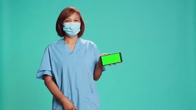 Nurse presenting explanatory healthcare tape on mock up chroma key green screen. Hospital employee holding phone in landscape mode, showing video, isolated over blue studio background