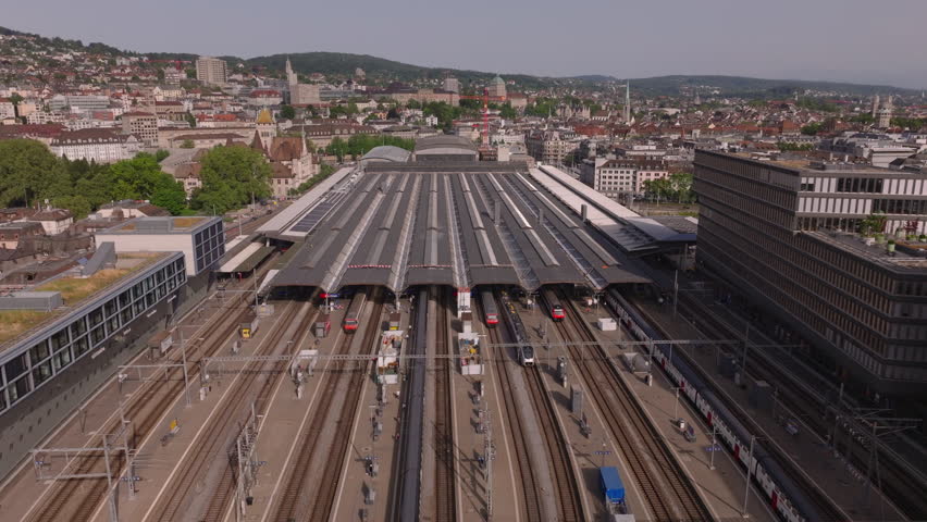 Forwards fly above main train station. Roofed platform and passenger trains on tracks. Aerial view of railway infrastructure in city. Zurich, Switzerland