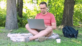 A man with glasses uses a laptop while sitting on a plaid on the grass in a park on a summer day. Remote work or online training