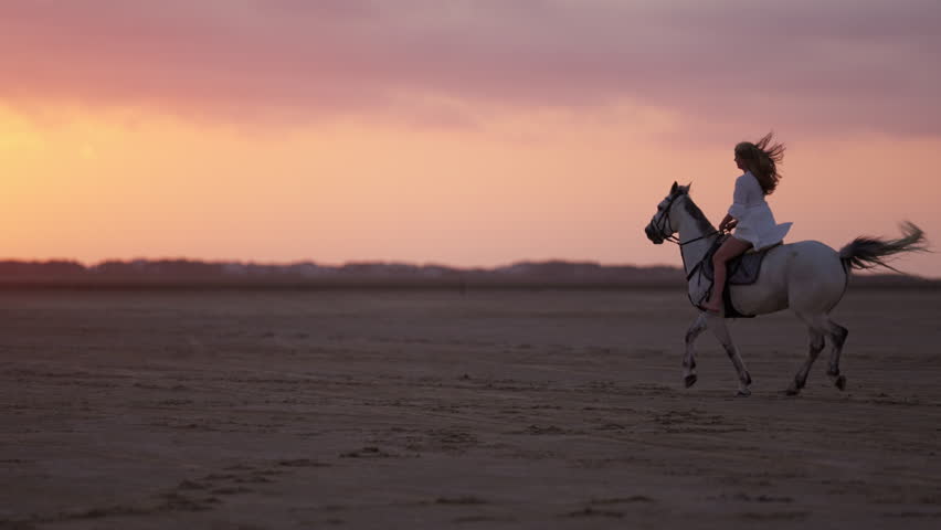 A wide shot of a long-haired girl in a white dress, riding a white horse on sandy soil in slow motion during an orange sunset