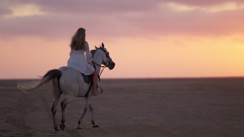A wide back view shot of a long-haired girl wearing a white dress, horse riding on a sandy soil during sunset