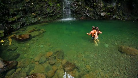 Happy Attractive Young Brunette Couple Swimming Towards the Base of a Giant Tropical Waterfall in a Blue Pond in Hawaii